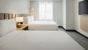 A hotel room with two beds, a nightstand with a lamp, a large window with sheer curtains, and a flat-screen TV mounted on the wall.