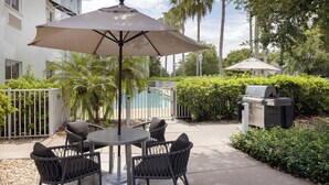 An outdoor patio with a round table and four chairs, an umbrella, a grill, and a pool in the background.