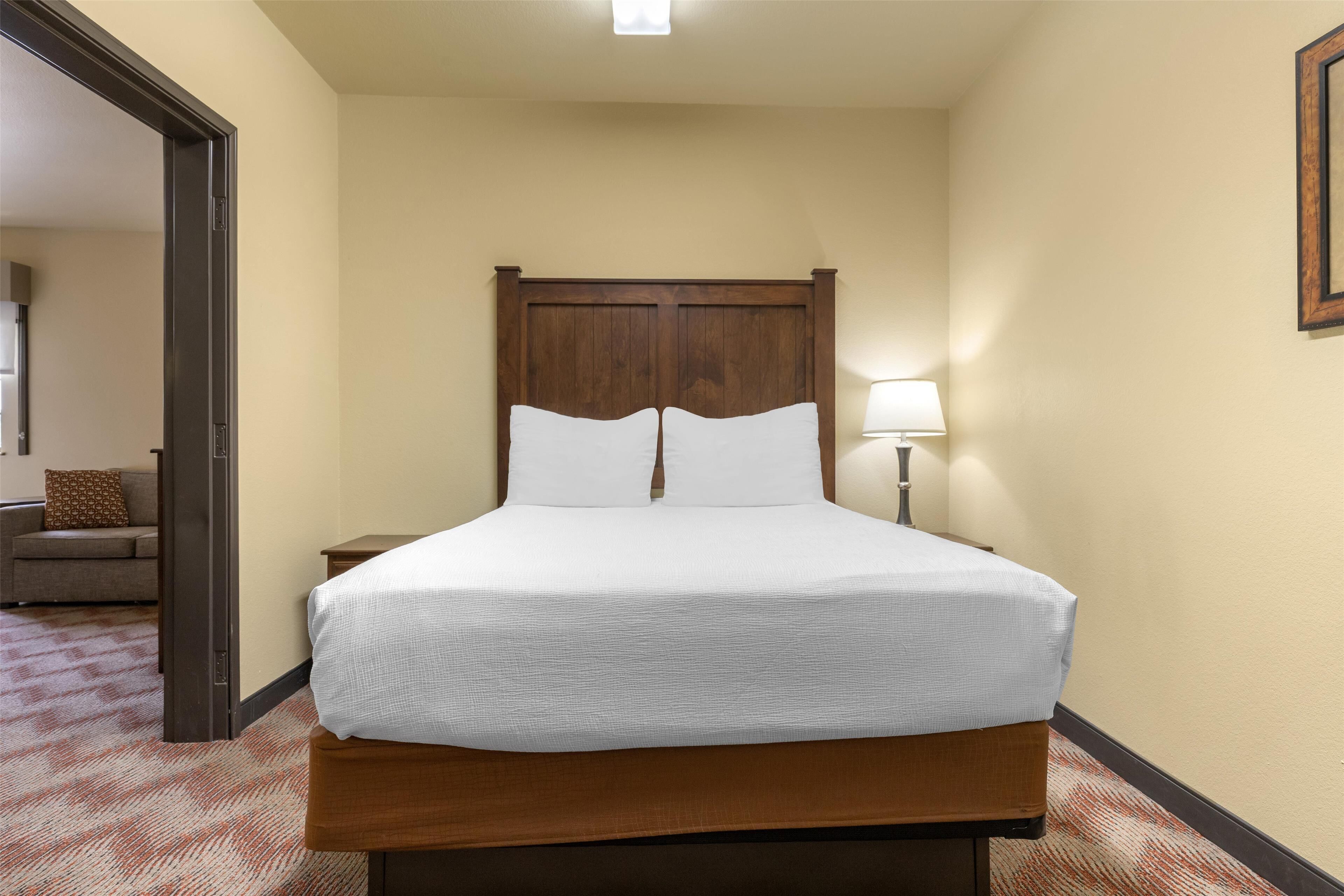 A hotel room with a wooden headboard, a single bed with white linens, a nightstand, a lamp, and a carpeted floor.