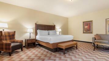 Una habitación de hotel con una cama, una silla, un banco, una mesita de noche, una lámpara, un sofá y un cuadro en la pared.