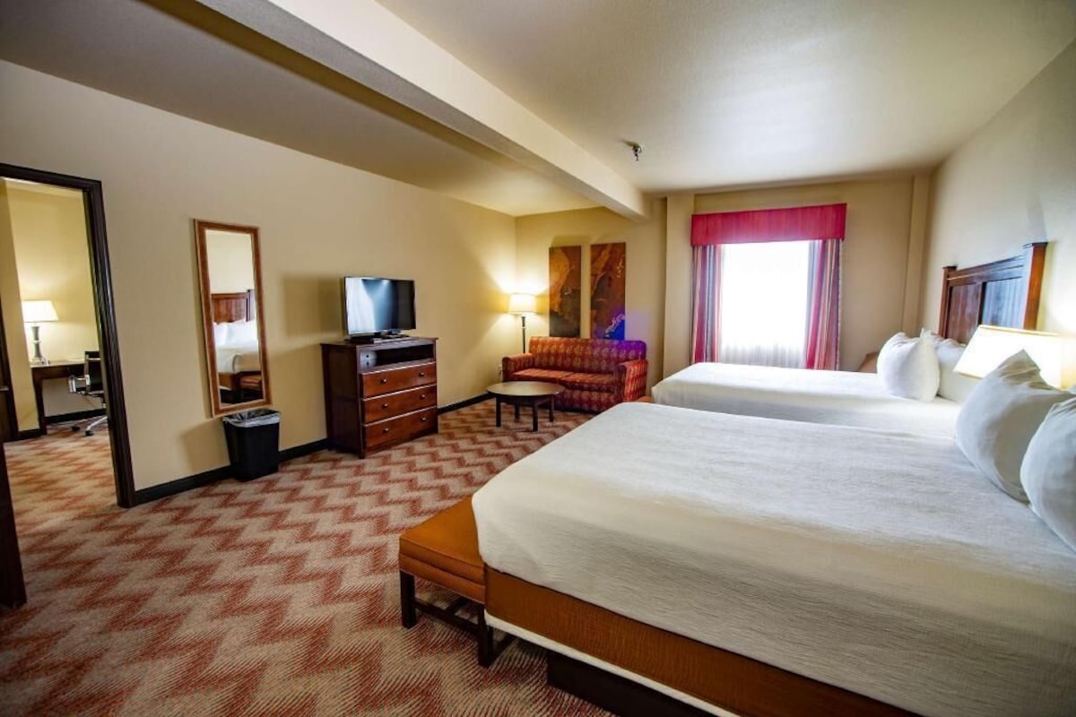Habitación de hotel con dos camas, televisión, escritorio con lámpara, silla y ventana con cortinas.