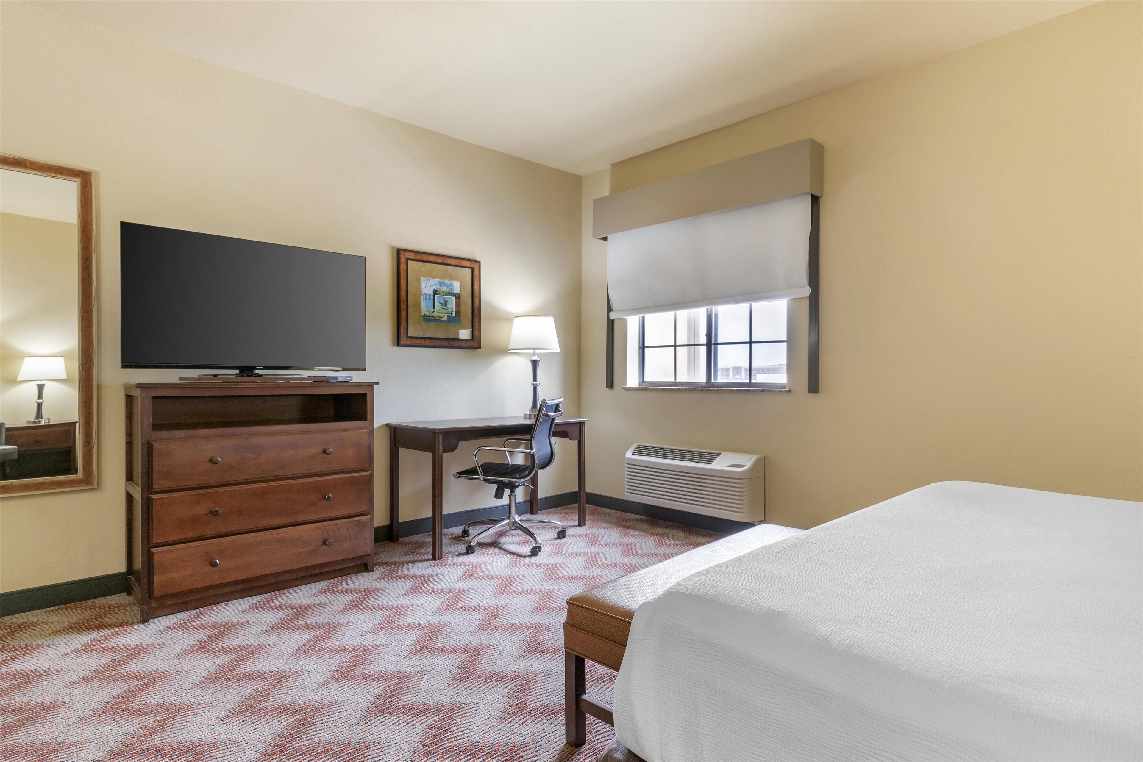 A hotel room with a bed, a desk with a chair, a dresser, a television, a window with a blind, and a wall-mounted air conditioning unit.