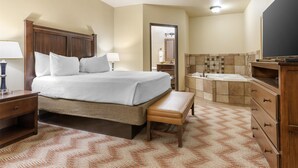 A hotel room with a large bed, a bedside table, a bench, a bathroom with a bathtub, and a TV. - Best Western Plus Cimarron Hotel & Suites (Stillwater)