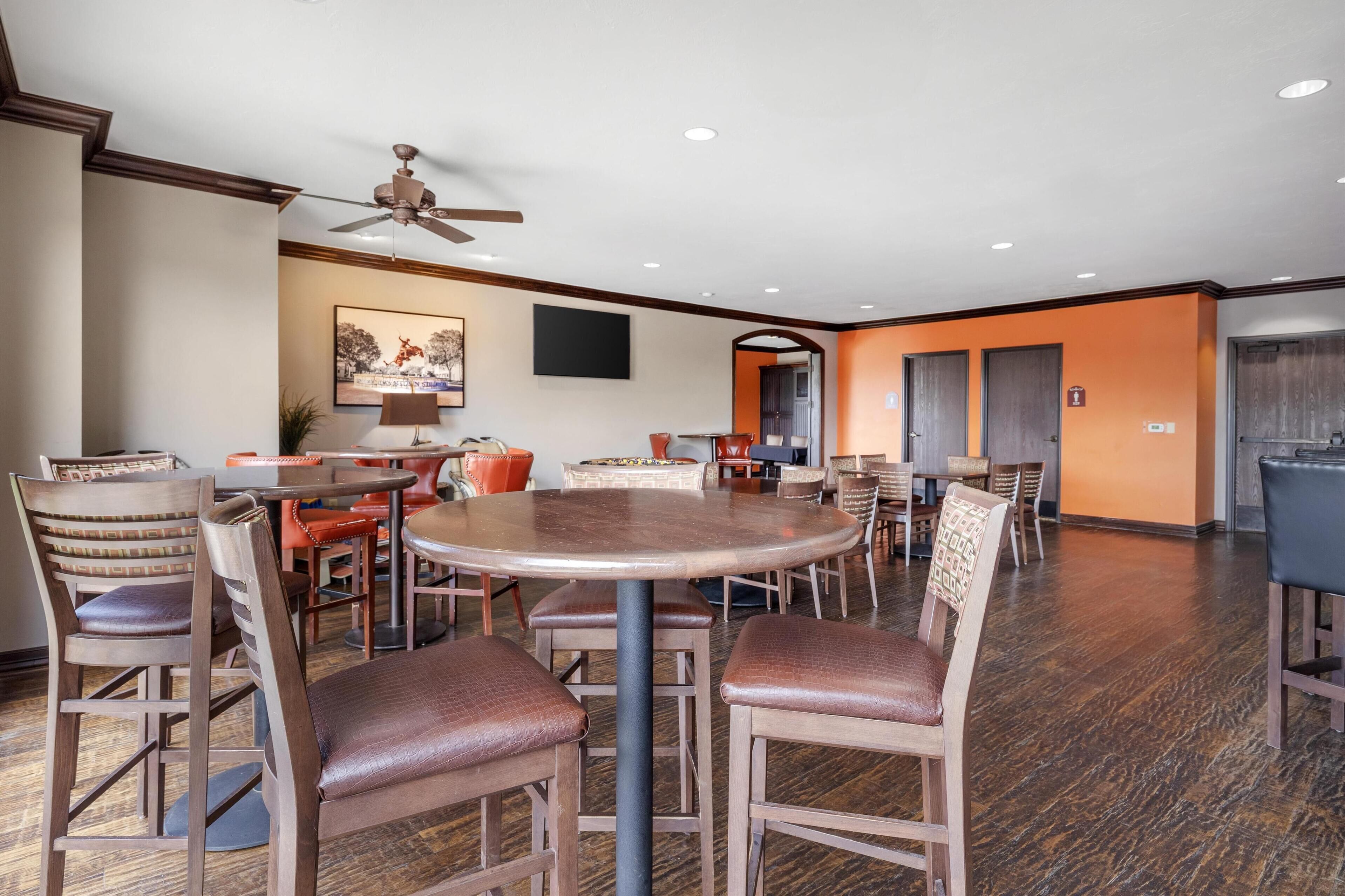 A spacious dining area with multiple round tables and wooden chairs, a ceiling fan, and a large painting on the wall.