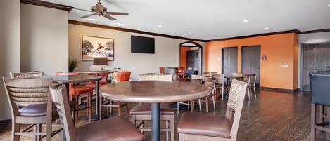A spacious dining area with multiple round tables and wooden chairs, a ceiling fan, and a large painting on the wall.