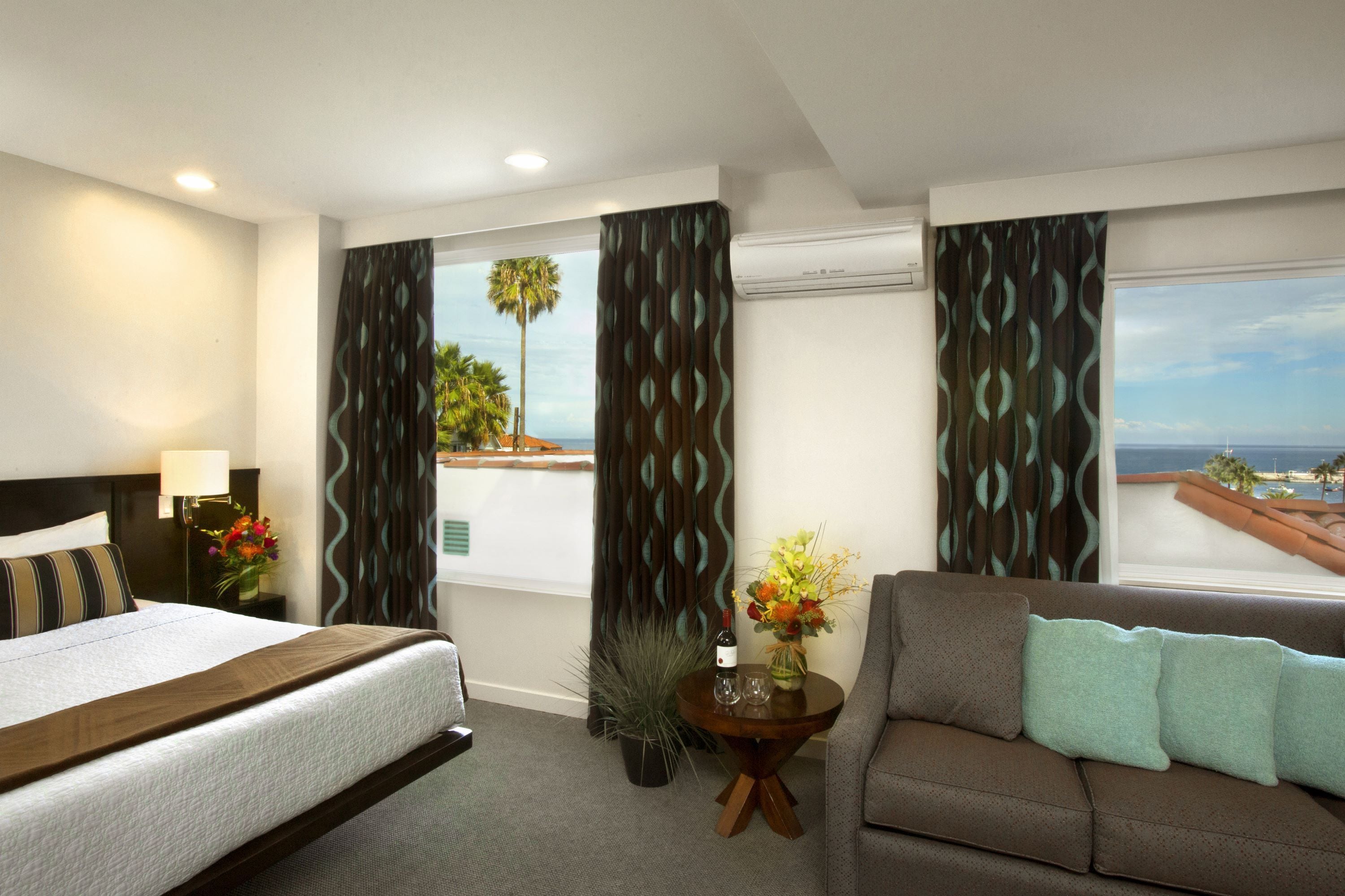 A hotel room with a bed, a sofa, a small table with a vase of flowers, and a view of the ocean through the windows.