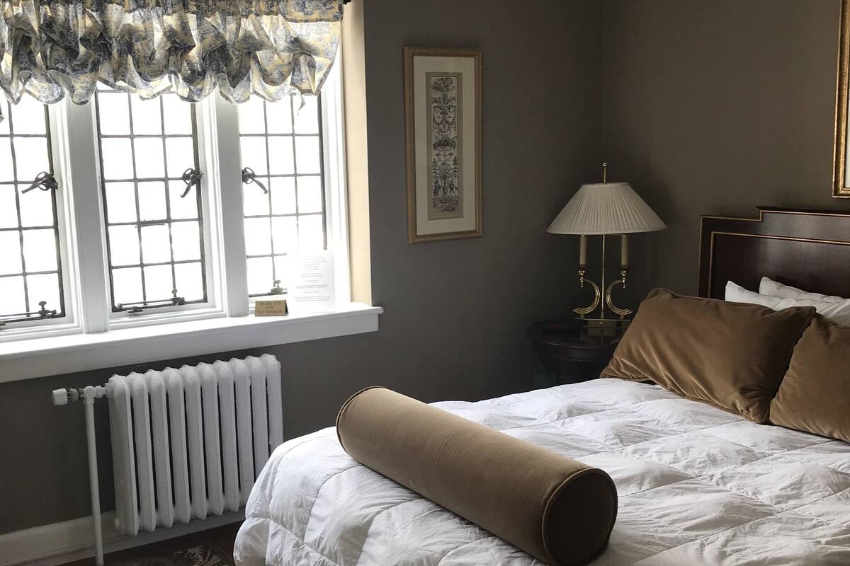 Une chambre d’hôtel avec un grand lit, un radiateur, une fenêtre donnant sur un beau paysage, une table de chevet avec une lampe et un tableau encadré au mur.