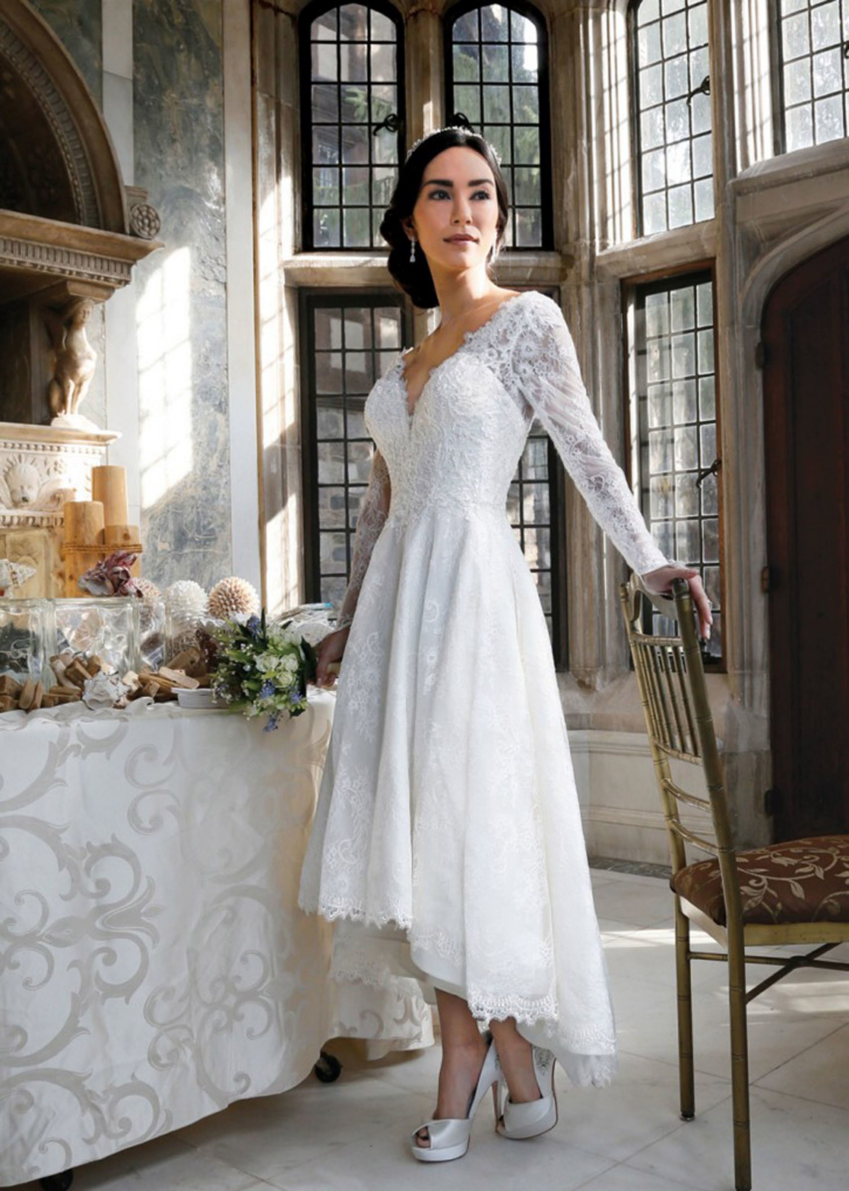 A person in a white lace dress standing in a room with large windows, a table with floral arrangements, and a chair.