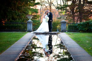 A couple standing on a reflective water path with a garden and trees in the background. - The Castle at Skylands Manor (Ringwood)