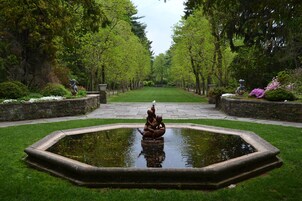 Um jardim com um lago central, rodeado por um caminho de pedra e vegetação exuberante.