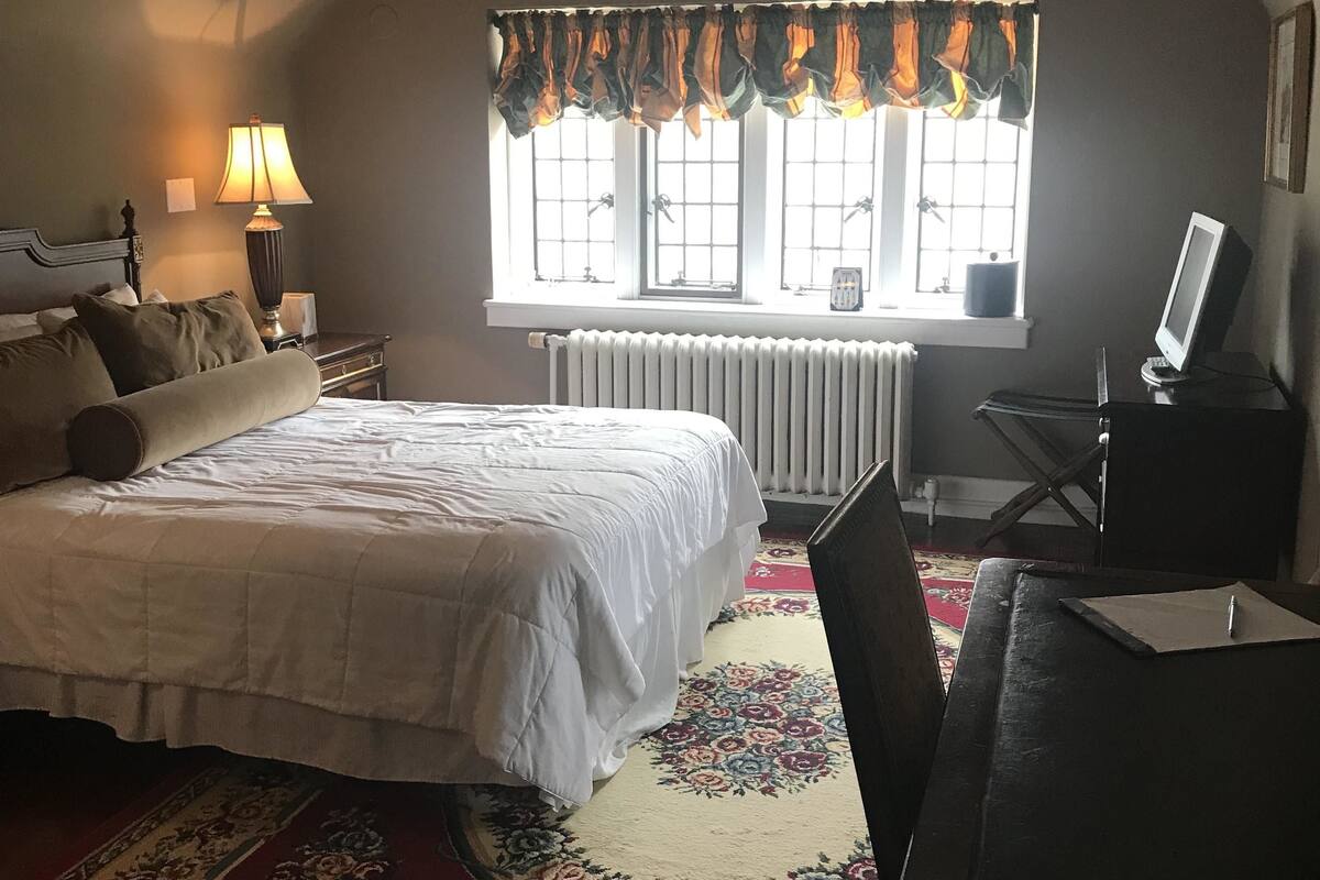 Une chambre à coucher avec un grand lit, un bureau avec un ordinateur, un radiateur et une fenêtre avec des rideaux.