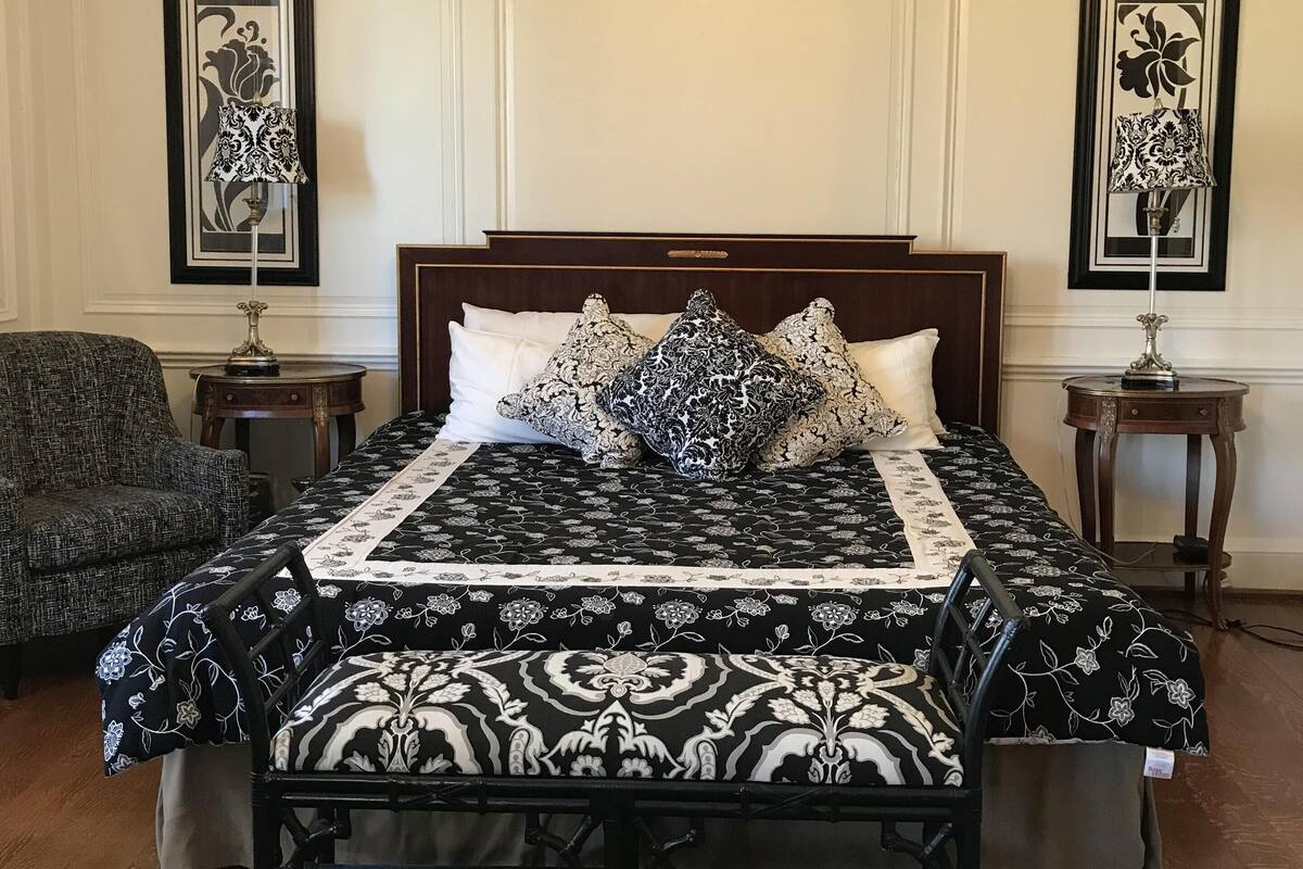 Une chambre à coucher avec un grand lit, deux tables de chevet, un lustre et deux tableaux représentant des motifs floraux, encadrés et accrochés aux murs.