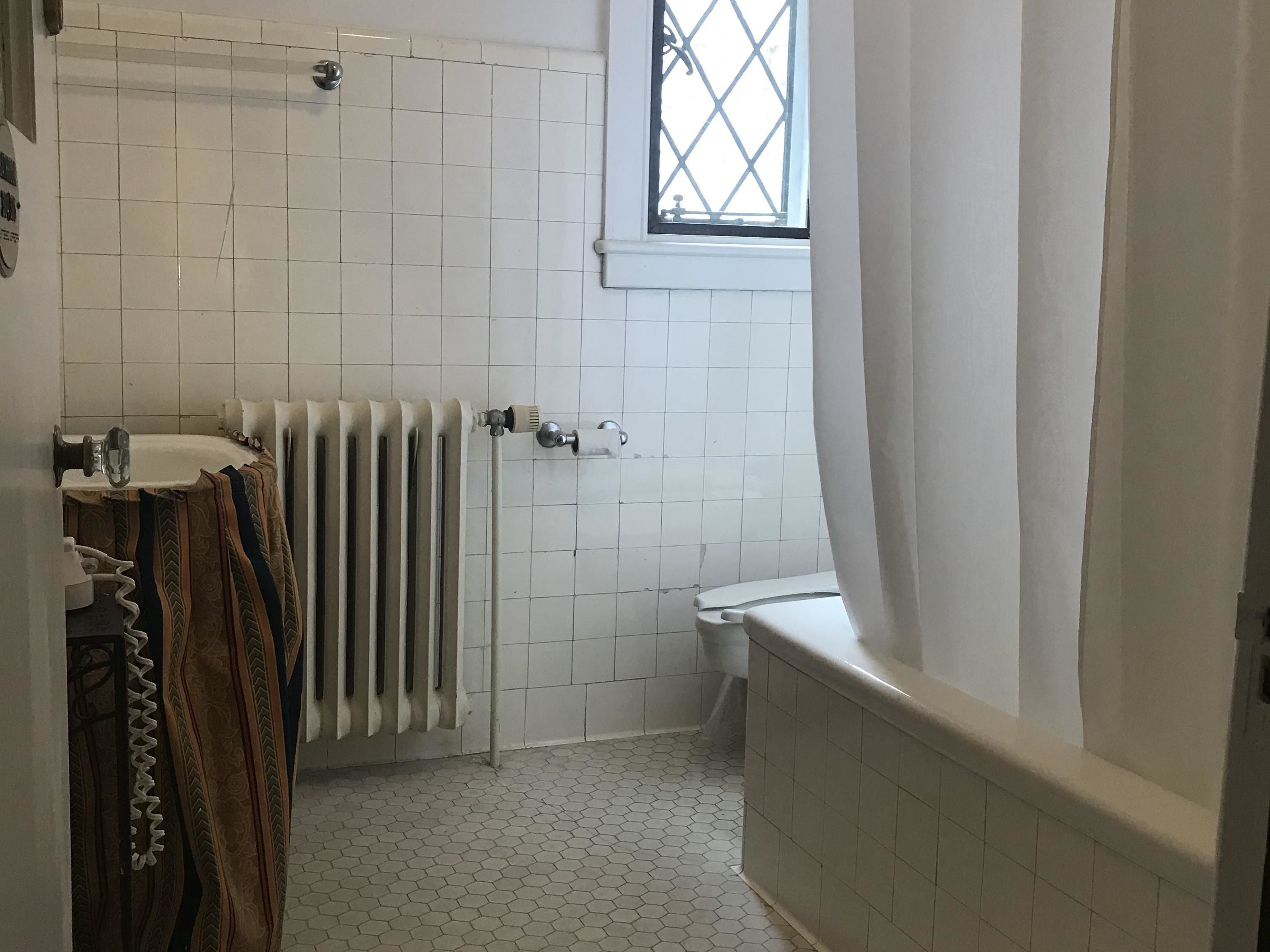 A bathroom with white tiled walls, a radiator, a toilet, and a window with a diamond pattern.