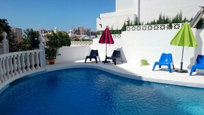 Une piscine à l’eau bleue, entourée de murs blancs et d’une clôture blanche. On y trouve deux chaises longues, l’une sous un parasol rouge et l’autre sous un parasol vert.
