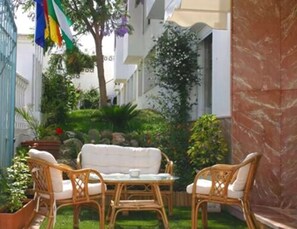 Un patio con mobiliario de mimbre y una mesita, rodeado de plantas y una bandera.