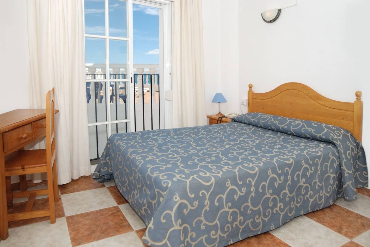 Une chambre à coucher comprenant un lit, un bureau en bois, une chaise, une table de chevet avec une lampe, et une vue sur l’extérieur par une fenêtre avec des rideaux.