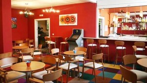Un restaurant aux murs rouges, avec de nombreuses tables et chaises, un bar où sont exposées des bouteilles, et un plafond agrémenté de luminaires.