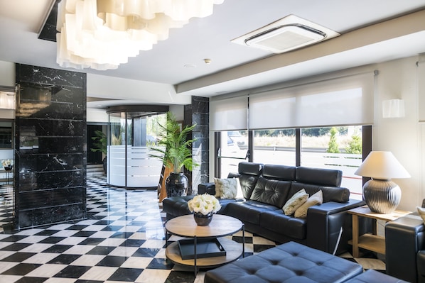 A modern lobby with a checkered floor, black leather sofas, a round coffee table with a vase of flowers, and a large window offering a view of the outdoors.