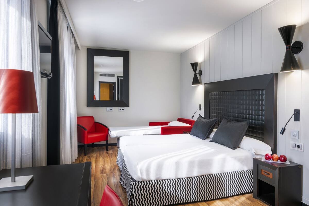 A modern hotel room with a large bed, a red chair, a desk, and a mirror.