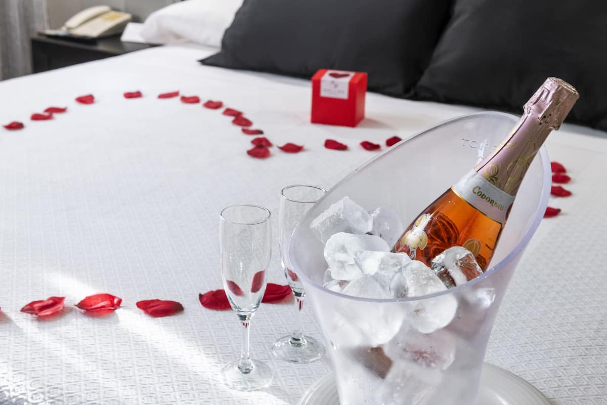 A bed with rose petals arranged in a heart shape, a bottle of champagne, and two glasses on a white tablecloth.