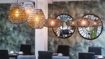A modern dining area with a tropical leaf-patterned wall, a round mirror, and three hanging woven light fixtures.