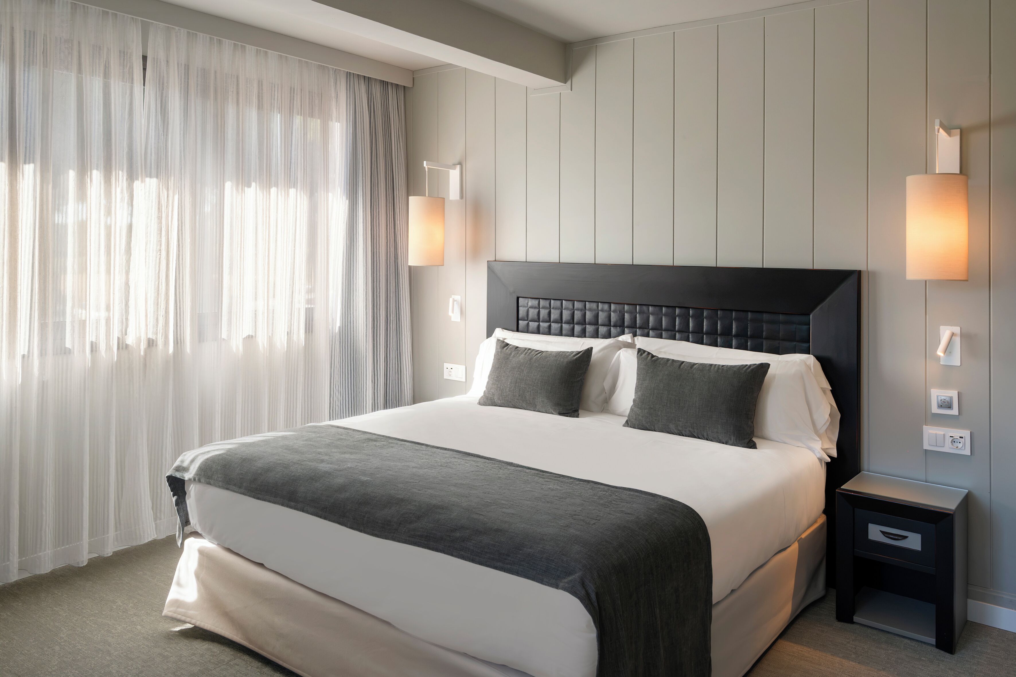 A hotel room with a large bed, two bedside lamps, a nightstand, and a window with sheer curtains.
