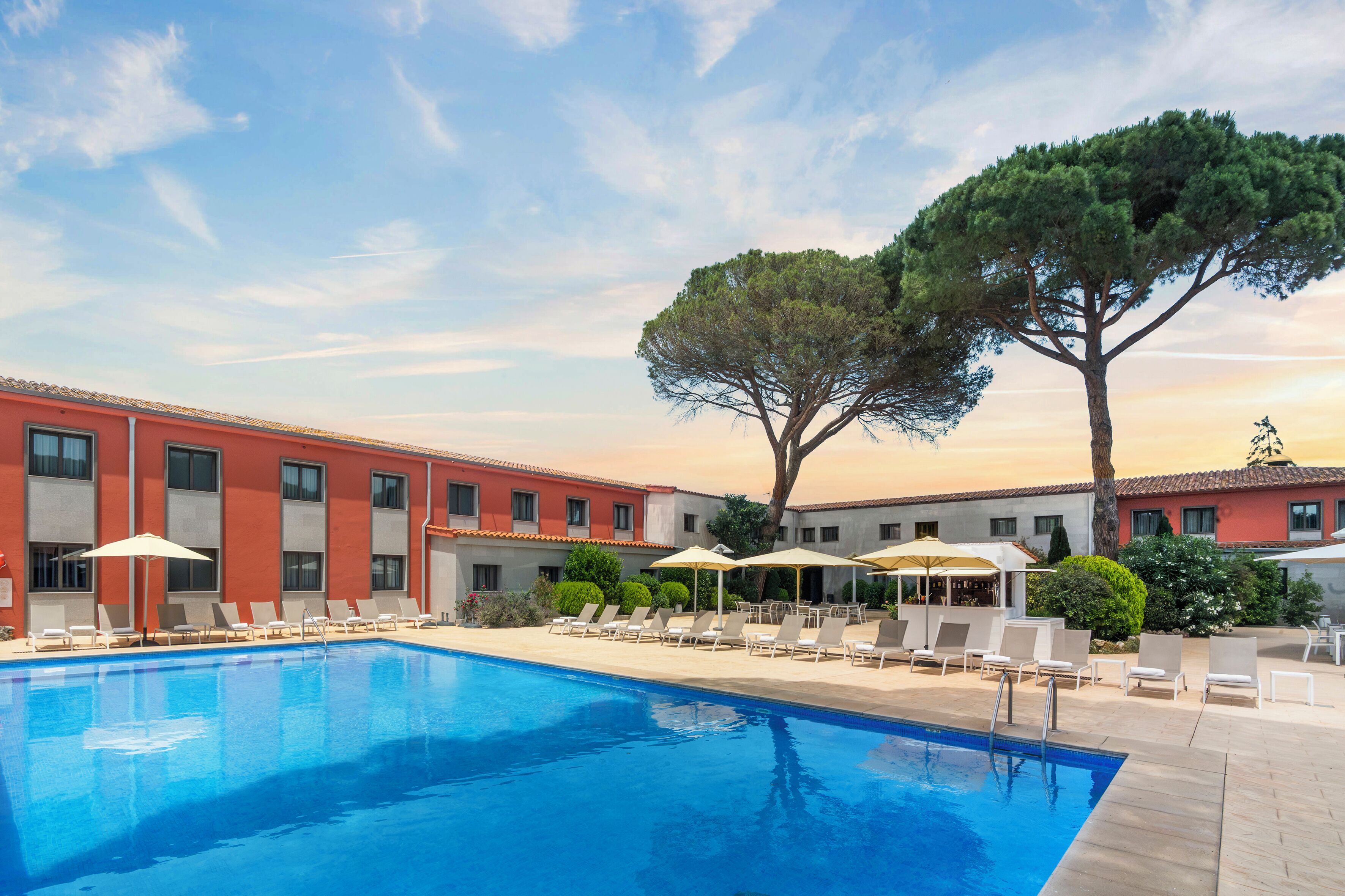 A swimming pool with lounge chairs and umbrellas, surrounded by a building with a tiled roof.