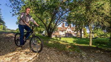 A man riding a bicycle on a gravel path with a large tree and a house in the background.