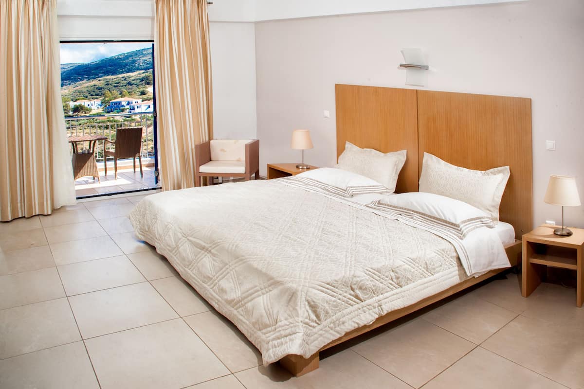 Habitación de hotel con una cama grande, dos mesitas de noche, una silla y un balcón con vistas al campo.