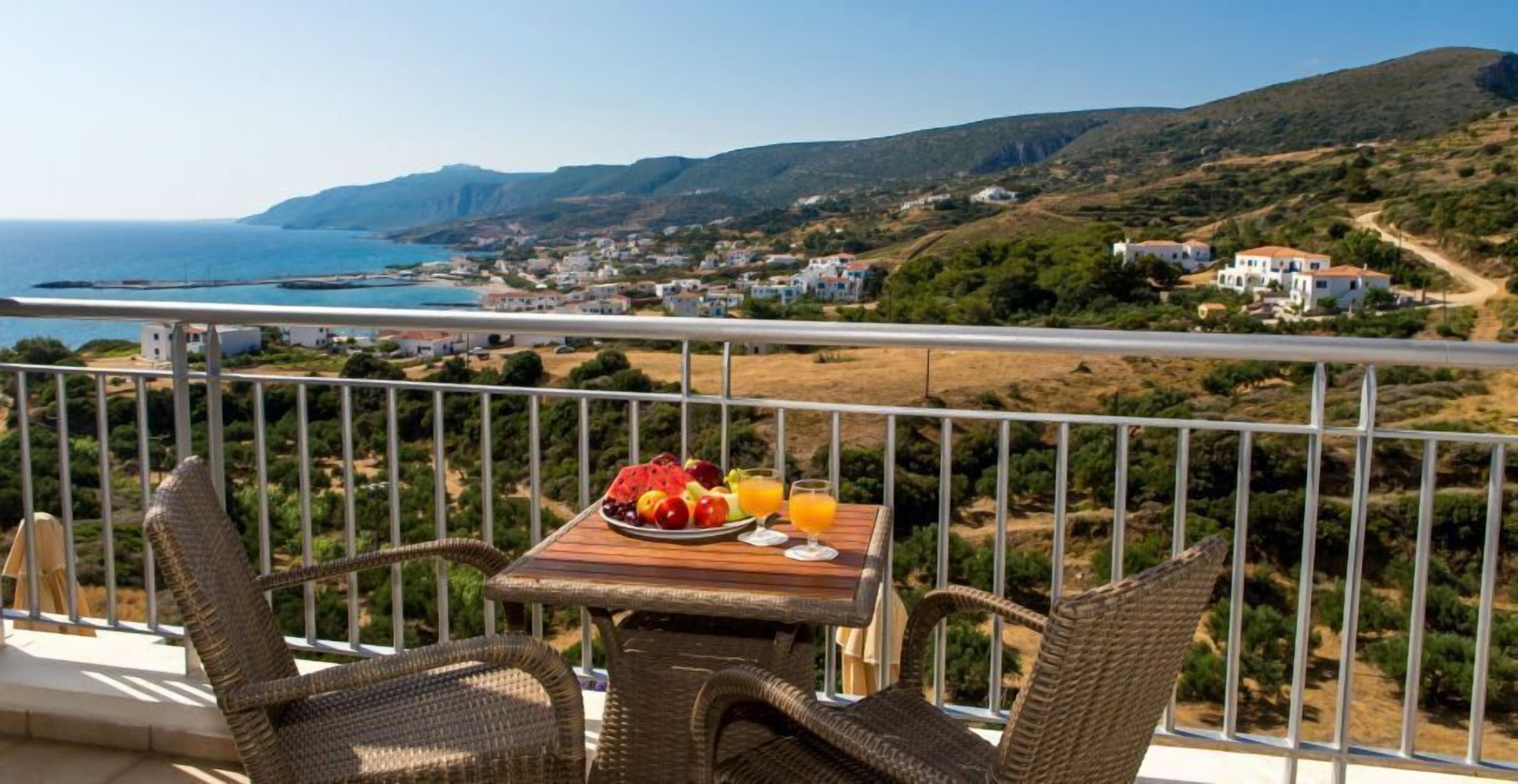 A balcony with wicker chairs and a table set with fruit and juice, overlooking a coastal town and hills.