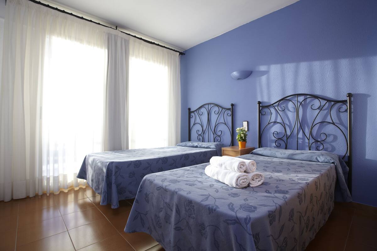 A hotel room with two beds, a window with white curtains, and a blue wall.