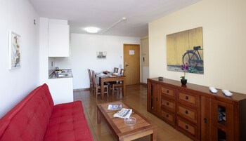 A compact living space with a red sofa, a wooden dining table with chairs, a kitchen area with a sink and cabinets, and a wooden cabinet with drawers.