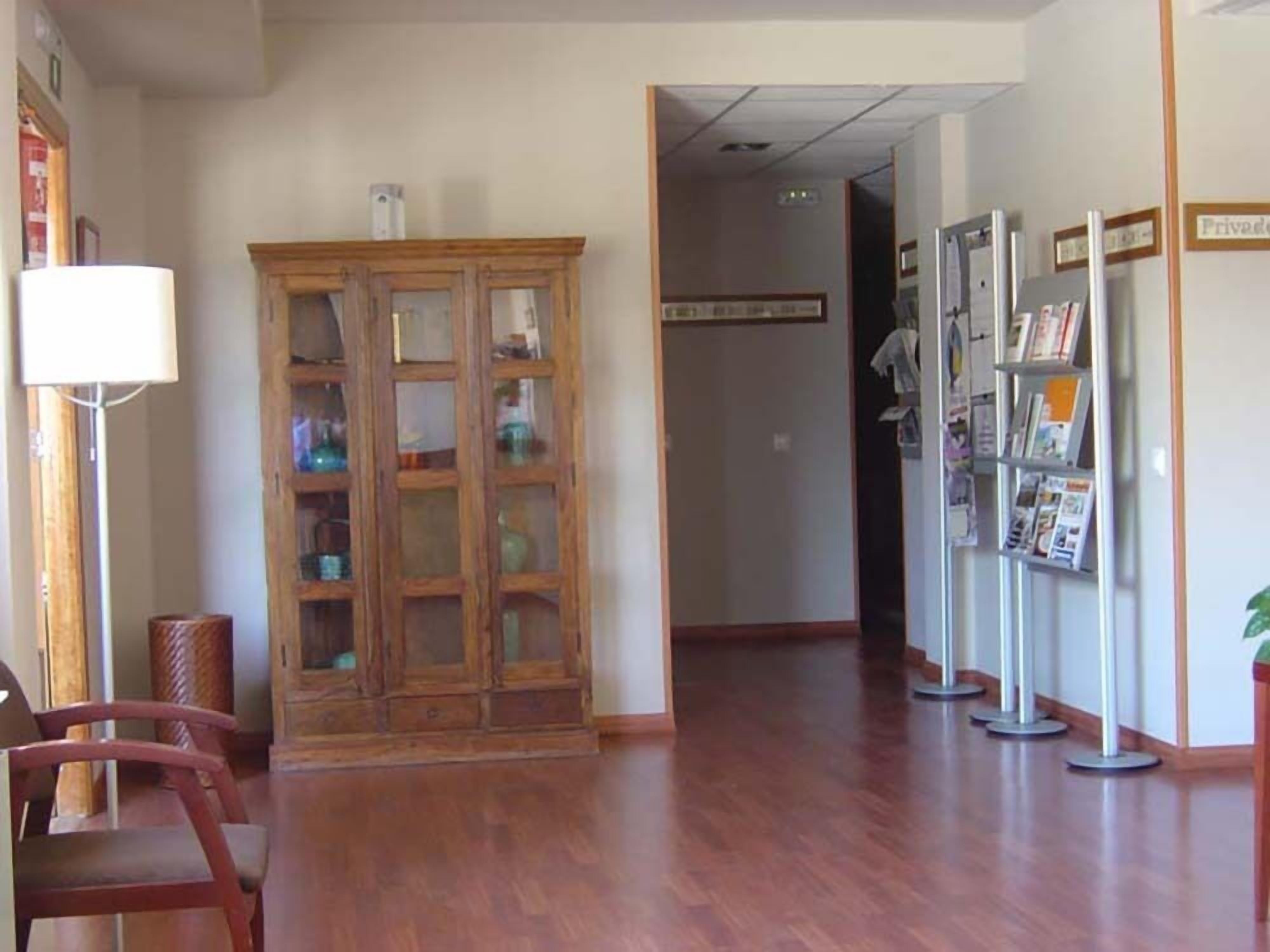A reception area with a wooden cabinet, a floor lamp, a chair, and a bookshelf with magazines.