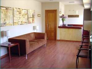 A room with a wooden floor, a brown sofa, a small table with a lamp, and a reception desk with a vase of flowers. - Hotel Midama (Chillaron de Cuenca)