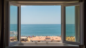 A view through a window of a beach with a clear sky, calm sea, and a sandy shore with umbrellas and people. - Pato Rojo (Punta Umbría)