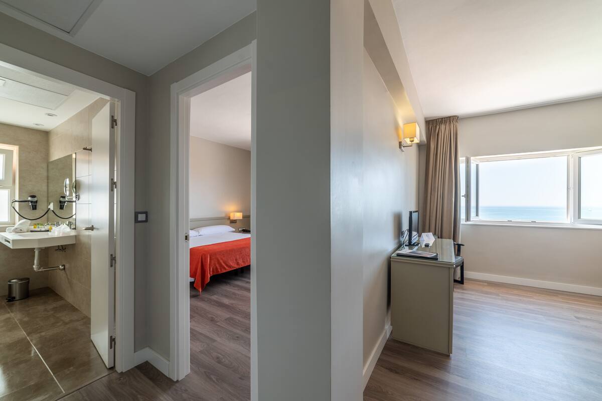 A hotel room with a bathroom, a bed with an orange bedspread, a desk with a lamp, and a window with a view of the sea.