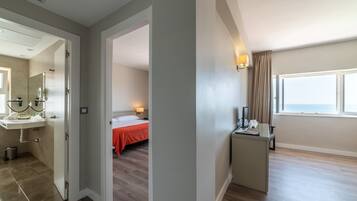 Una habitación de hotel con baño, una cama con colcha naranja, un escritorio con lámpara y ventana con vista al mar.