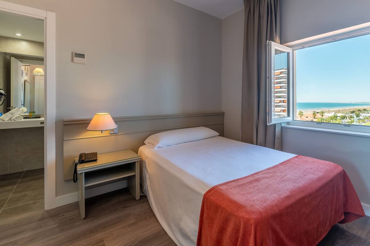 A hotel room with a bed, a nightstand with a lamp, a mirror, and a window with a view of the beach.