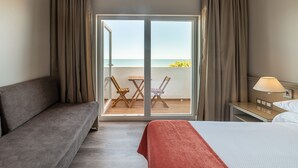 A hotel room with a bed, a sofa, a table with chairs, a lamp, and a view of the sea through a glass door. - Pato Rojo (Punta Umbría)