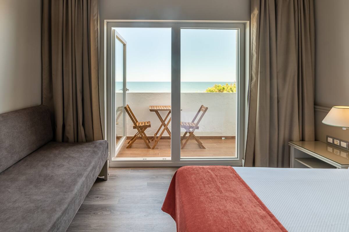 A hotel room with a bed, a sofa, a table with chairs, a lamp, and a view of the sea through a glass door.