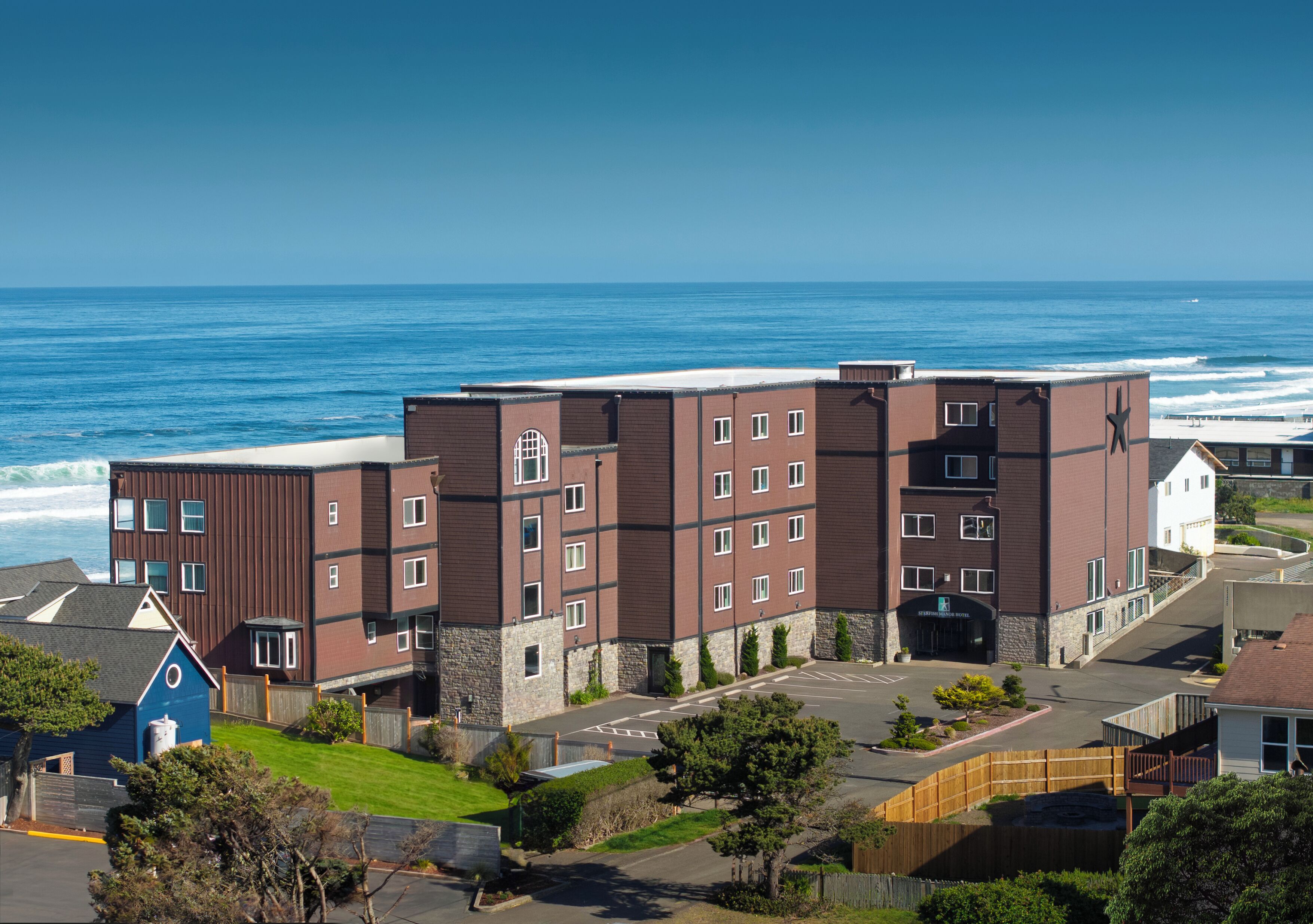 A multi-story hotel with a parking lot, a view of the ocean, and a small building with a blue roof.