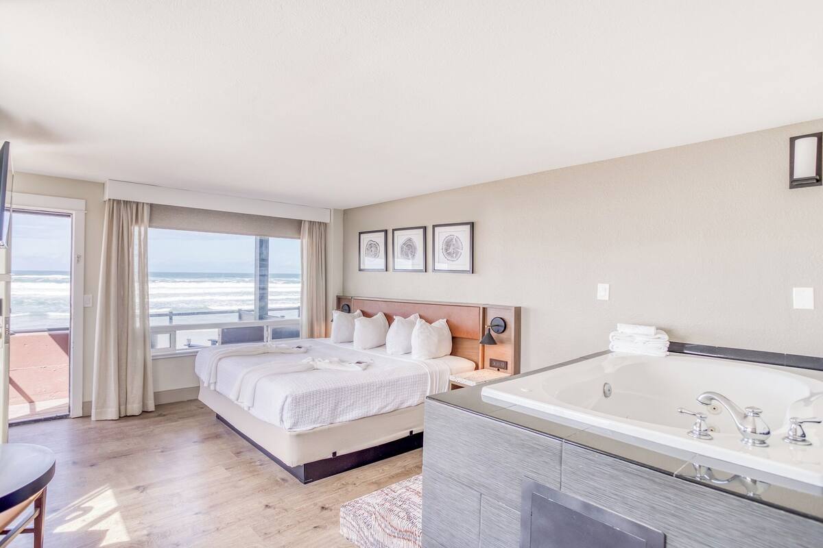 A hotel room with a large bed, a bathroom with a bathtub, and a view of the ocean.