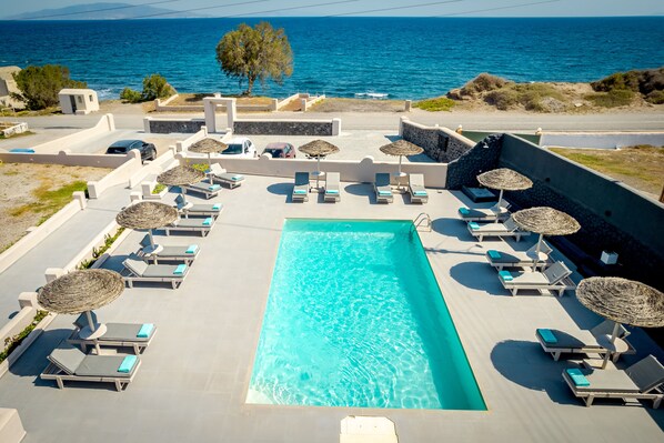 A pool area with lounge chairs and umbrellas, a clear blue pool, and a view of the sea. - En Plo Boutique Suites (Santorini)