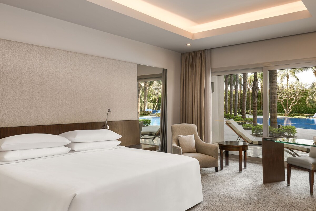 Habitación de hotel con una cama grande, una silla, una mesita y vistas a una piscina.