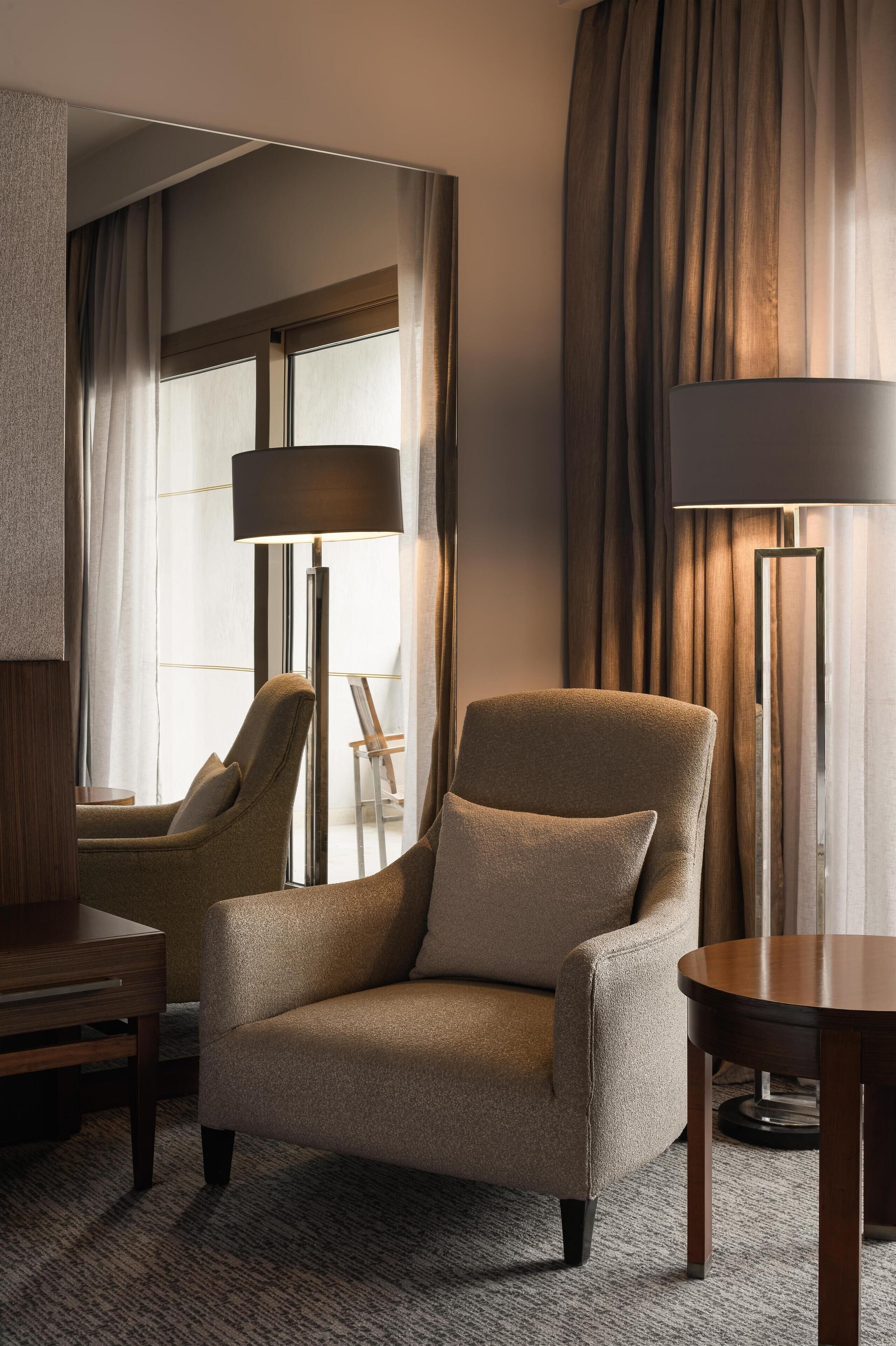 Habitación de hotel con un sillón color beige, una mesa redonda de madera, una lámpara de pie y vistas al exterior a través de cortinas.