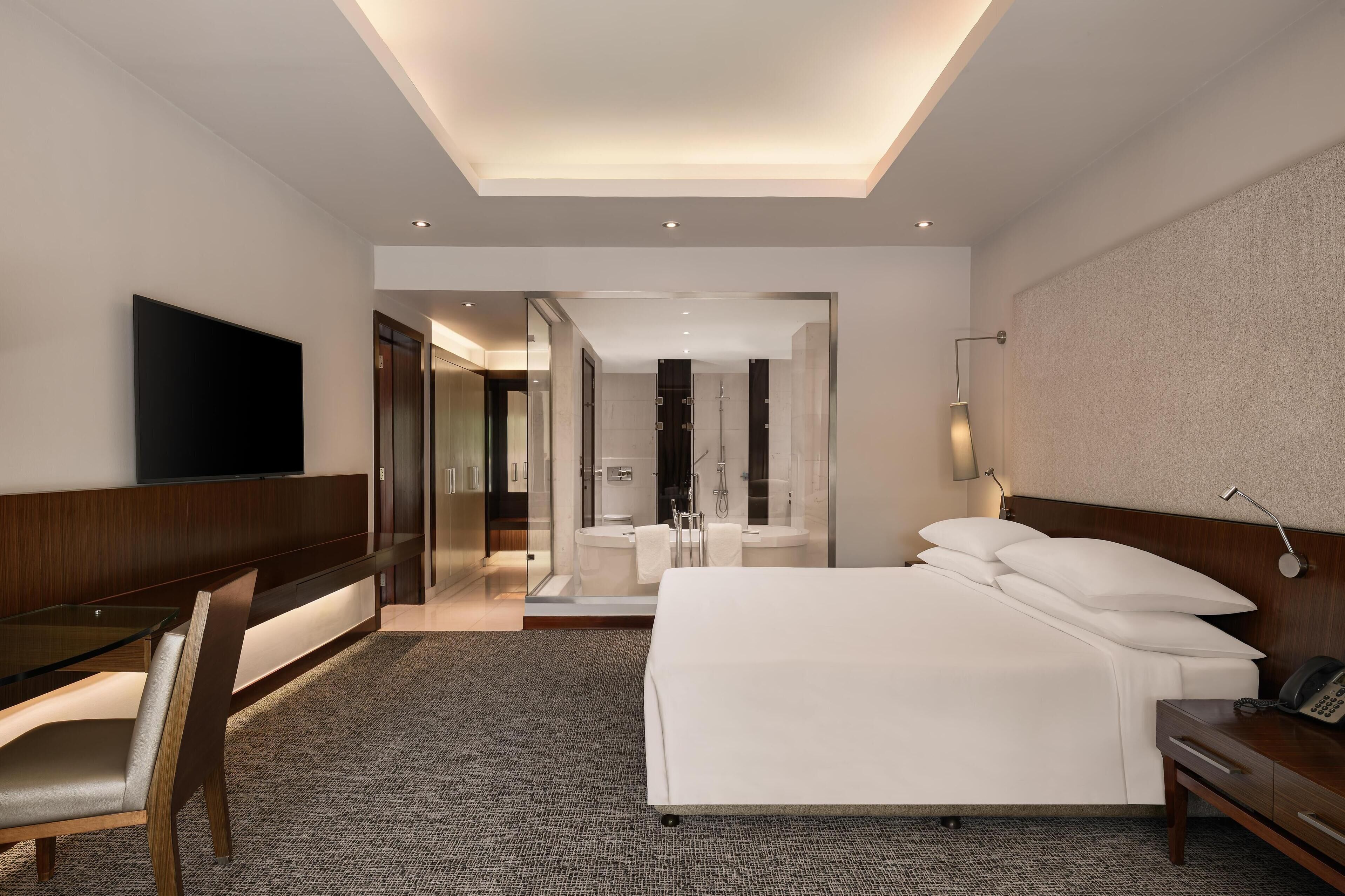 A modern hotel room with a large bed, a desk, a TV, and a bathroom visible through a glass door.