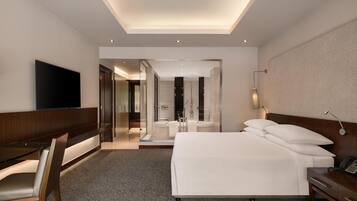 A modern hotel room with a large bed, a desk, a TV, and a bathroom visible through a glass door.