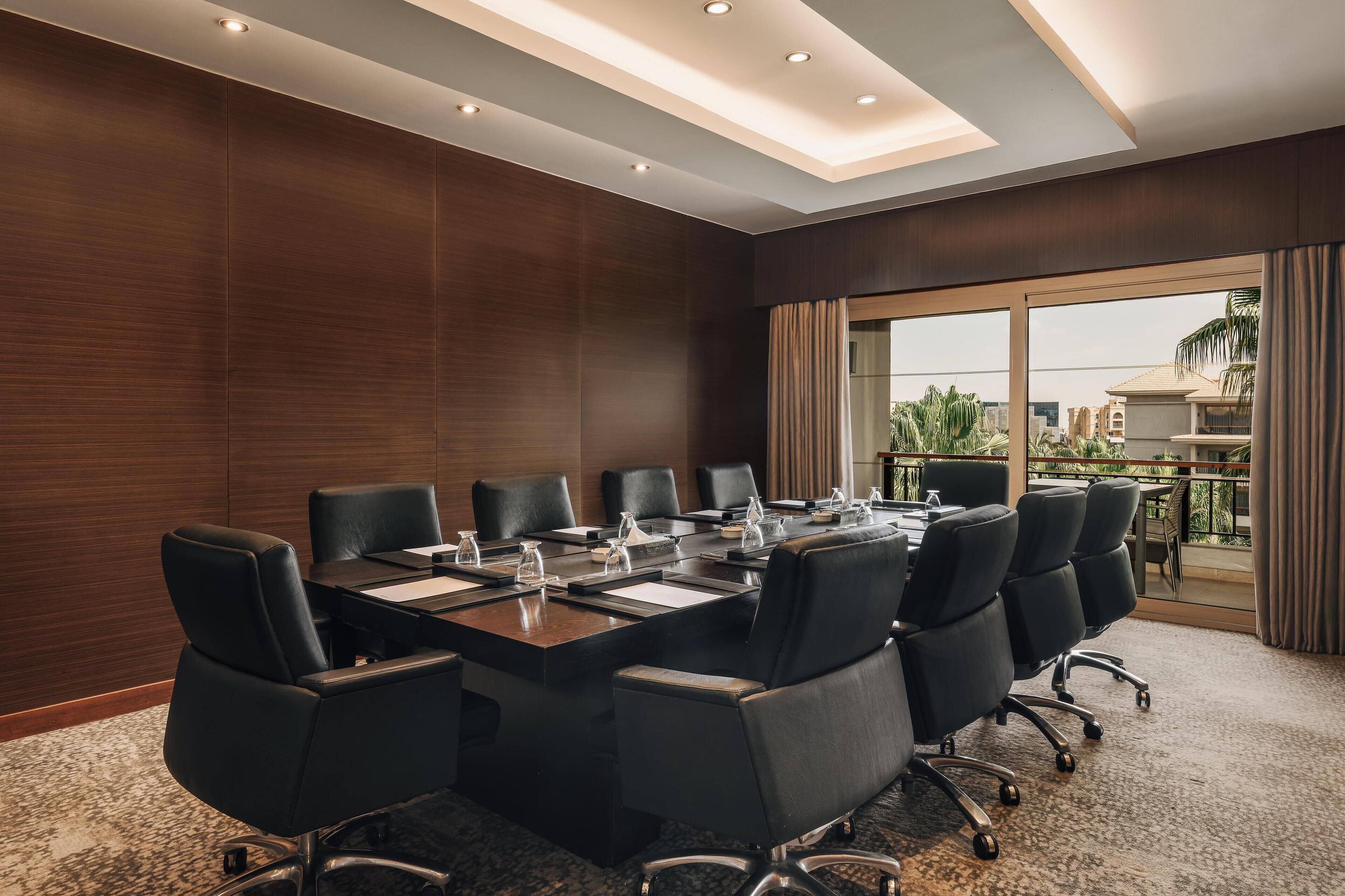 Une salle de conférence avec une longue table, des chaises noires et une vue sur les palmiers, visible à travers de grandes fenêtres.