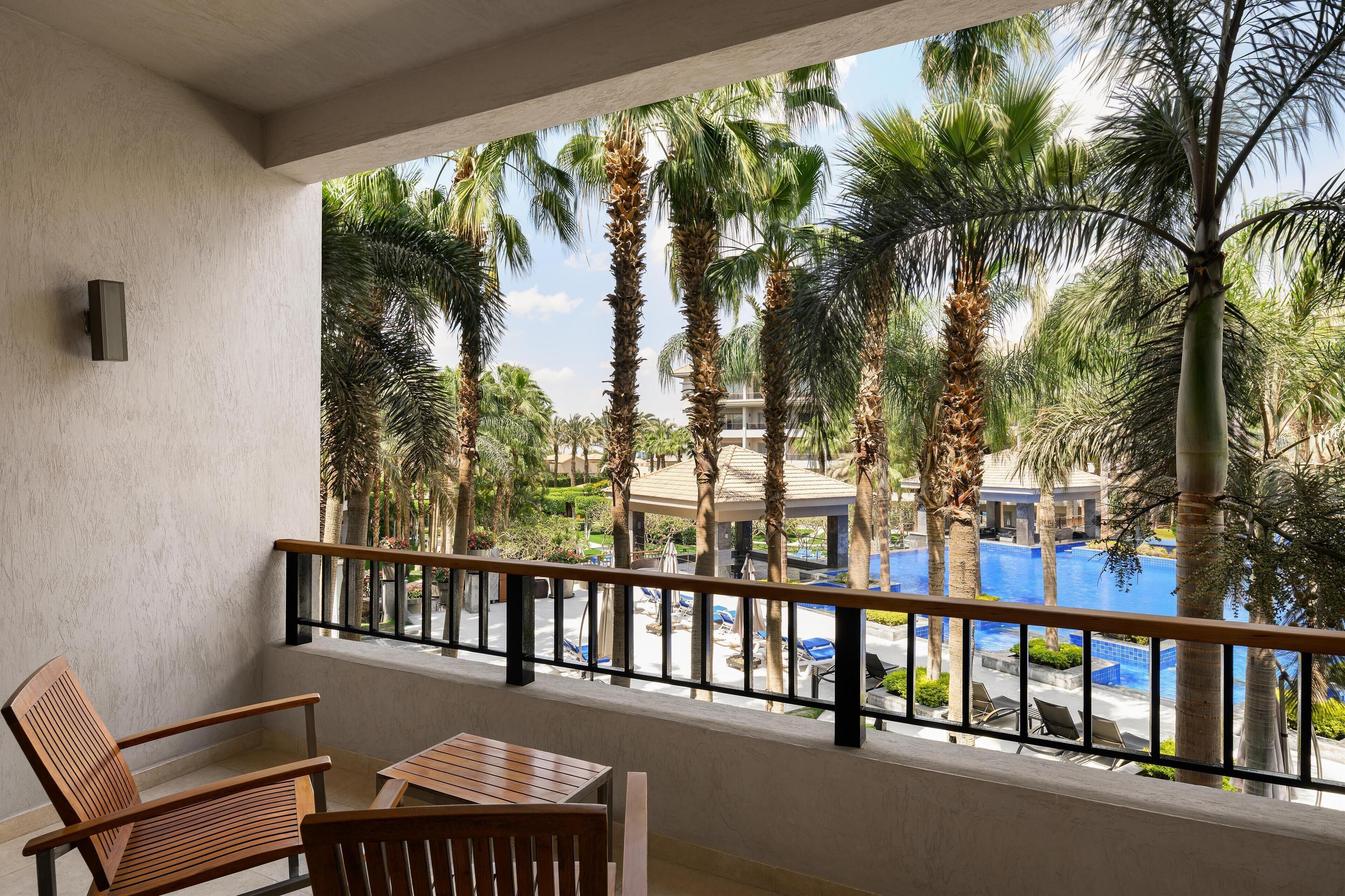 A balcony with wooden furniture, a pool, and palm trees.