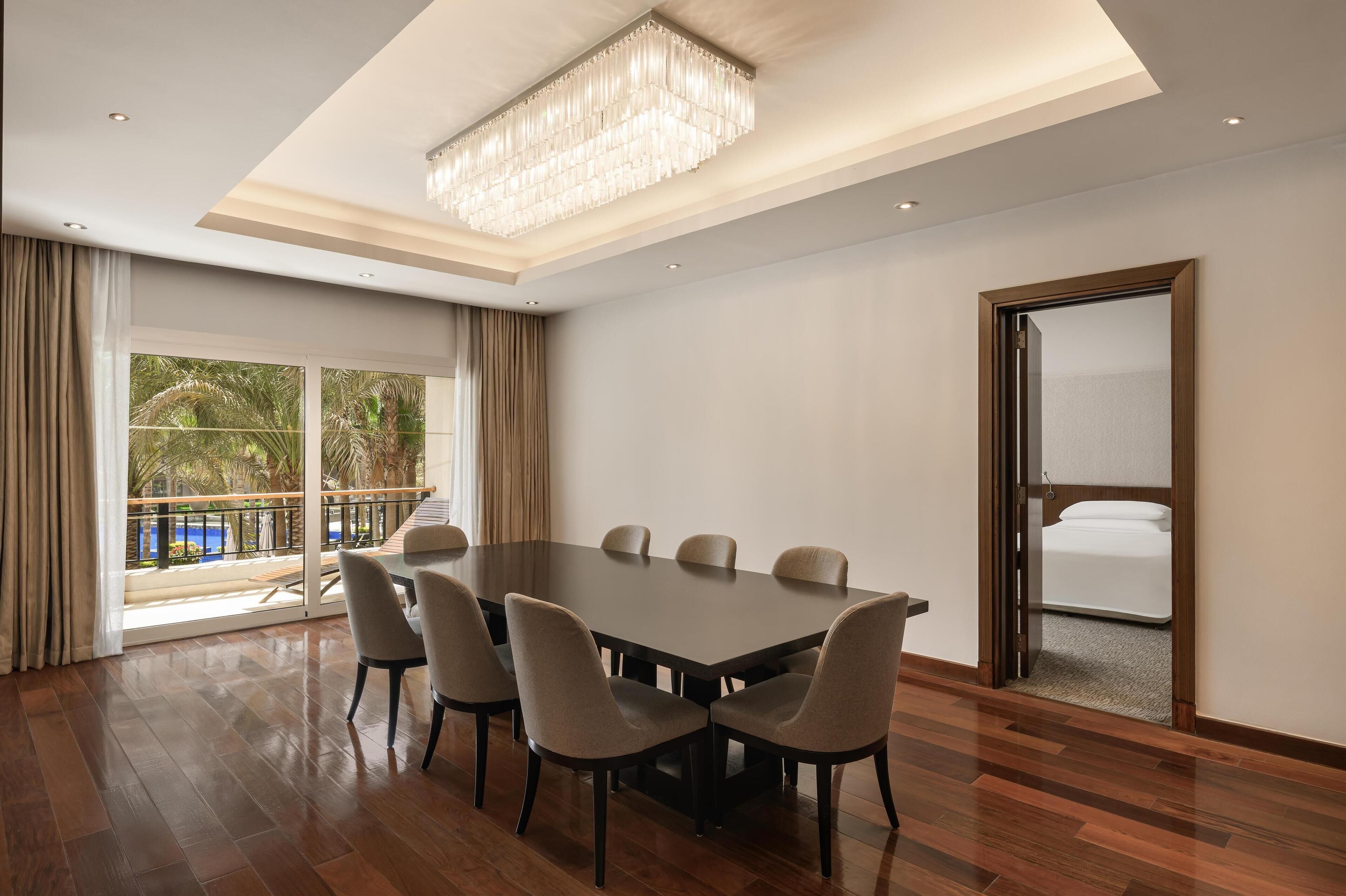 A spacious room with a large dining table, comfortable chairs, a chandelier, and a view of a pool through large windows.